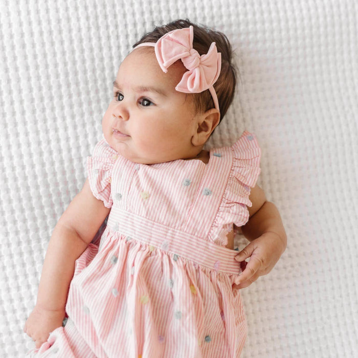 Baby wearing a pink dress and headband with a bow, lying on a textured white surface.