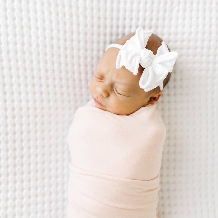 Newborn baby swaddled in pink with a white bow headband on a textured white background