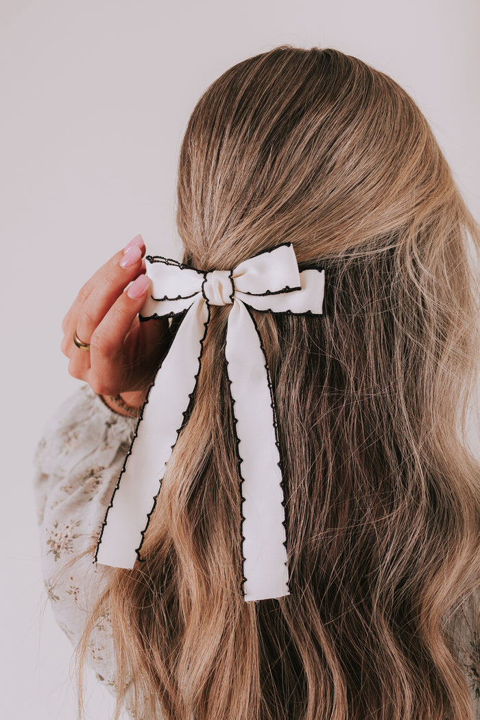 Person with long wavy hair wearing a decorative white bow in the hair against a neutral background