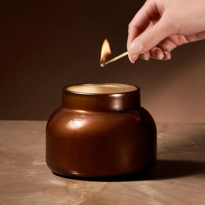 Brown glass container candle with a lit match in scent chestnut embers by capri blue.