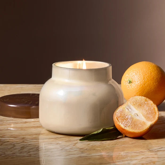 White pearlescent glass candle with brown glass lid with gold accents next to sugared orange in scent volcano by capri blue.