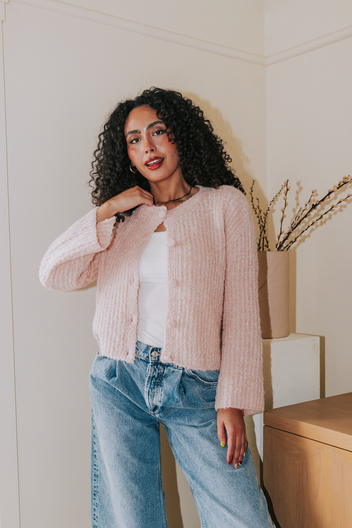 Woman wearing a light pink cardigan over a white top and blue jeans, standing in a room with a wooden cabinet and plant.