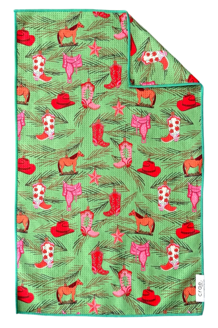Green microfiber hand towel with pine sprigs, horse ornaments, and pink and red cowboy boots, hats, and saddles.