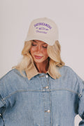 Woman wearing a beige cap with text and a denim jacket on a plain background