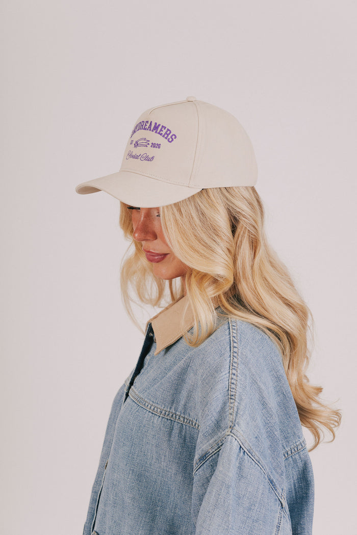 Woman wearing a beige cap with text and a denim jacket on a plain background