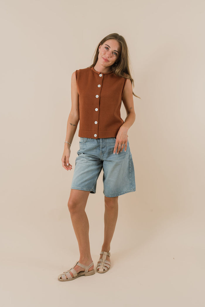 Woman wearing a brown sleeveless shirt with white buttons and light blue shorts on a beige background