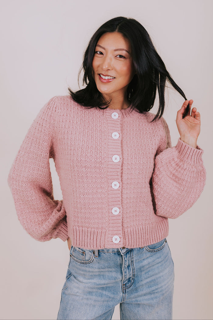 Woman wearing a pink knitted cardigan and blue jeans against a plain background