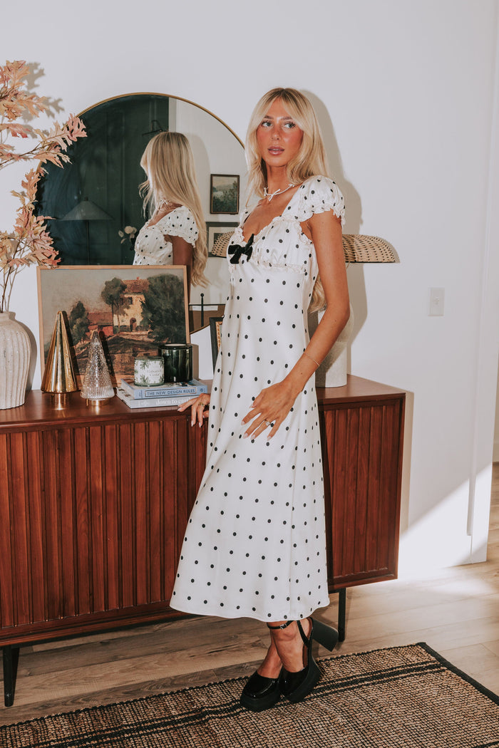 Woman in a white polka dot dress standing in a room with a wooden sideboard and decorative items.