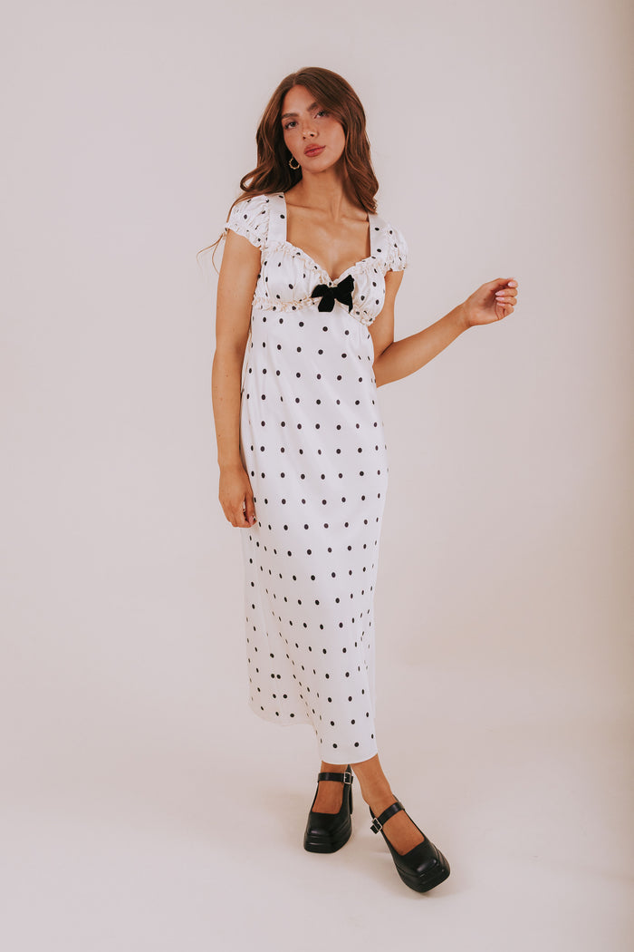 Woman wearing a white dress with black polka dots and a black bow on a plain background