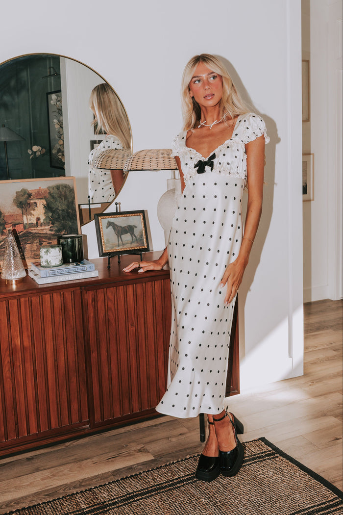 Woman in a white polka dot dress standing in a room with wooden furniture and decor.