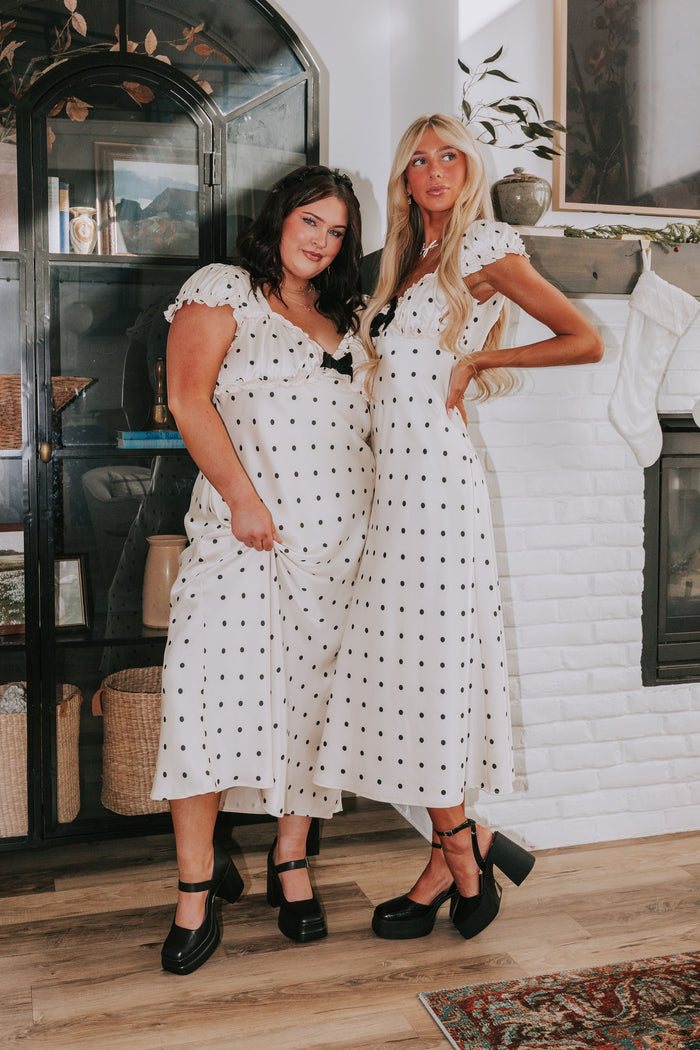 Two women in matching polka dot dresses standing in a room with a decorative archway and fireplace.