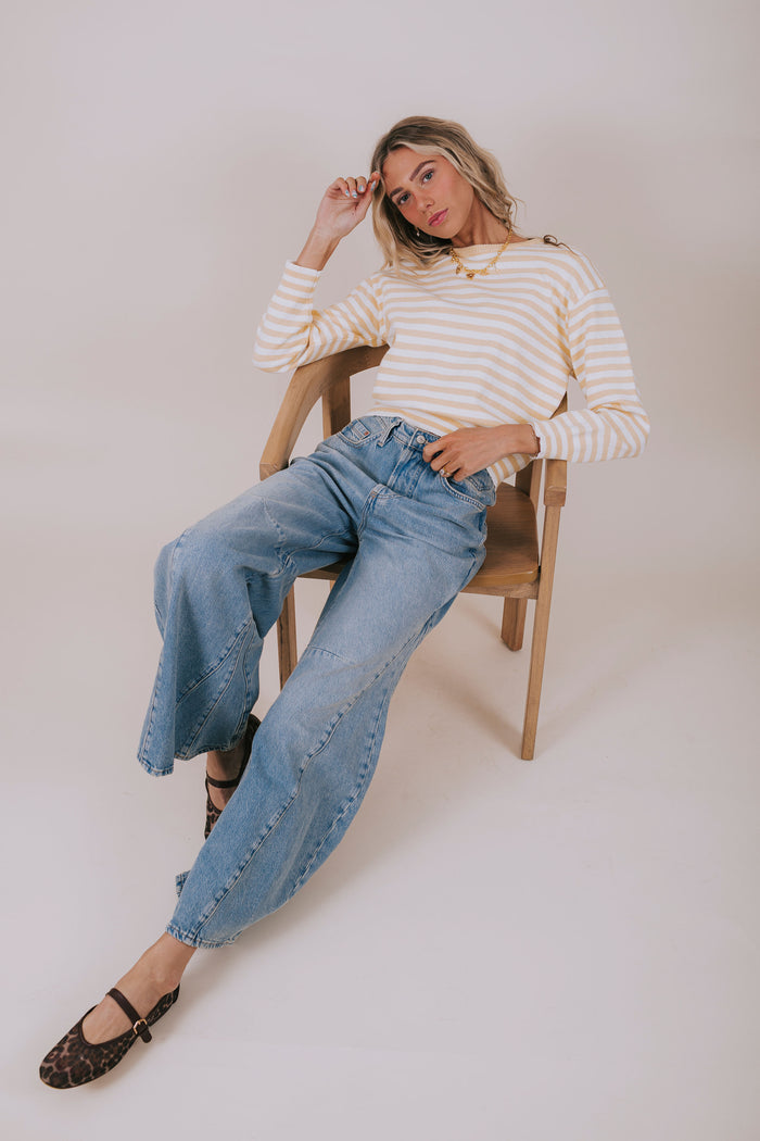 Woman sitting on a chair wearing a striped sweater and blue jeans against a plain background