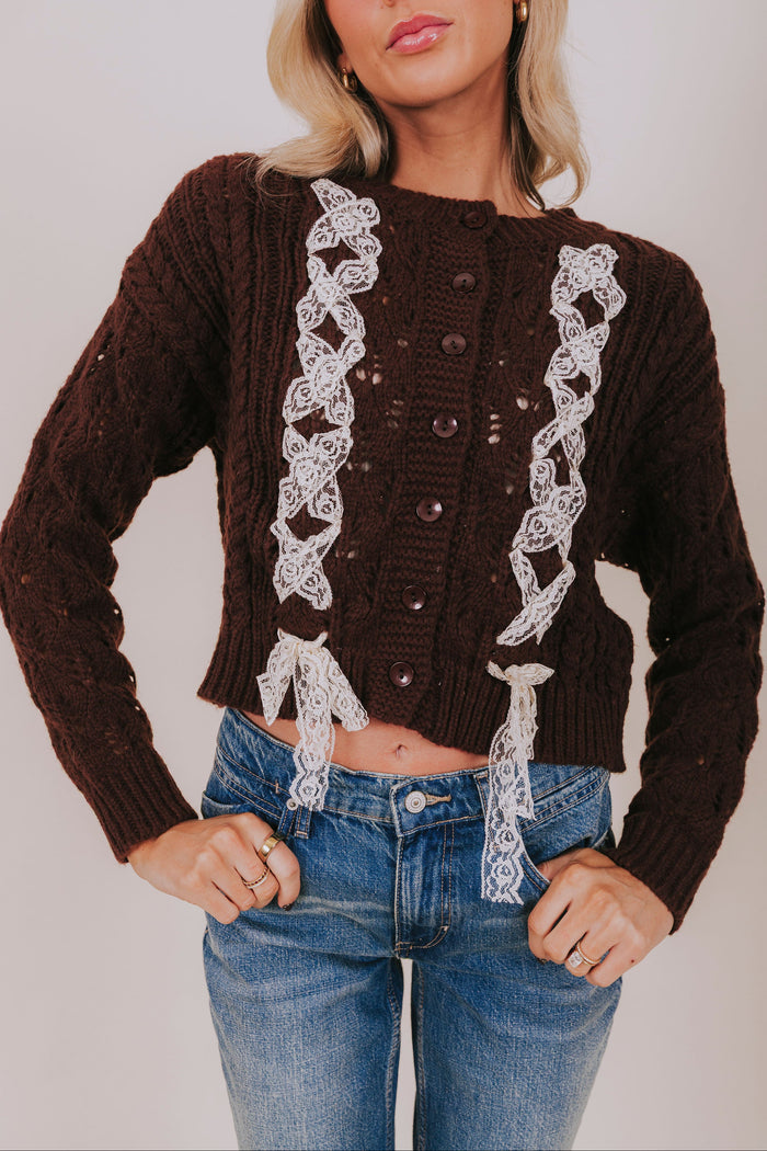 Woman wearing a brown sweater with lace details and blue jeans against a plain background