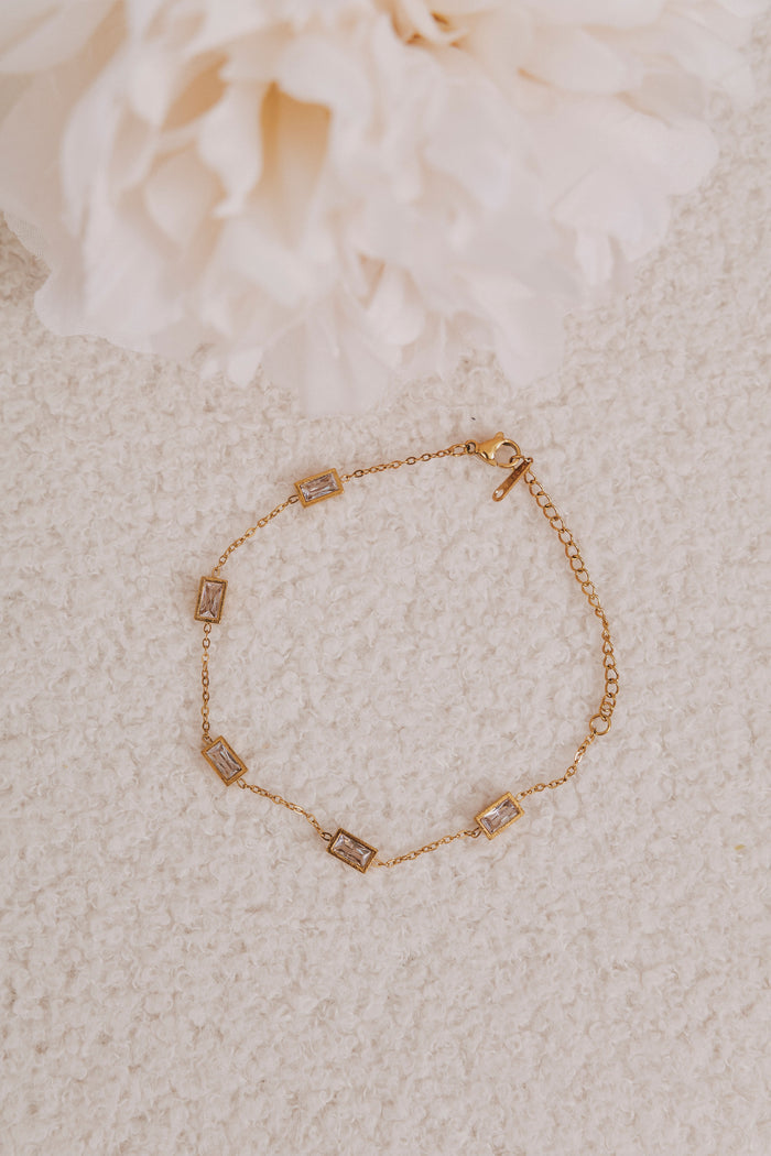 Gold bracelet with small rectangular charms on a textured beige surface