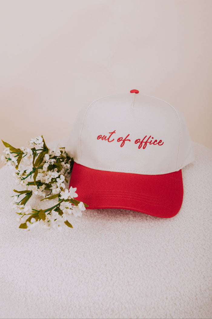 White cap with red brim and 'out of office' text, placed on a white surface with flowers.