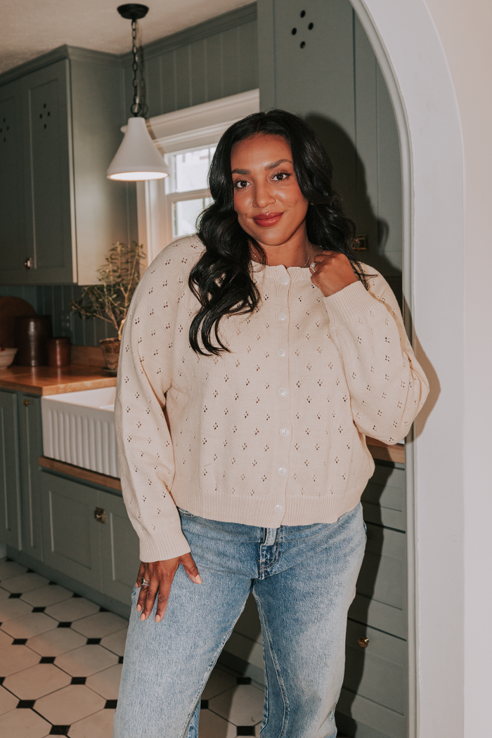 Woman standing in a kitchen wearing a beige sweater and blue jeans.