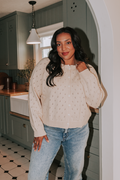 Woman standing in a kitchen wearing a beige sweater and blue jeans.
