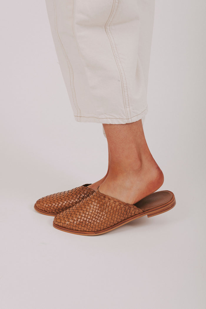 Brown woven slippers worn with light-colored pants on a white background