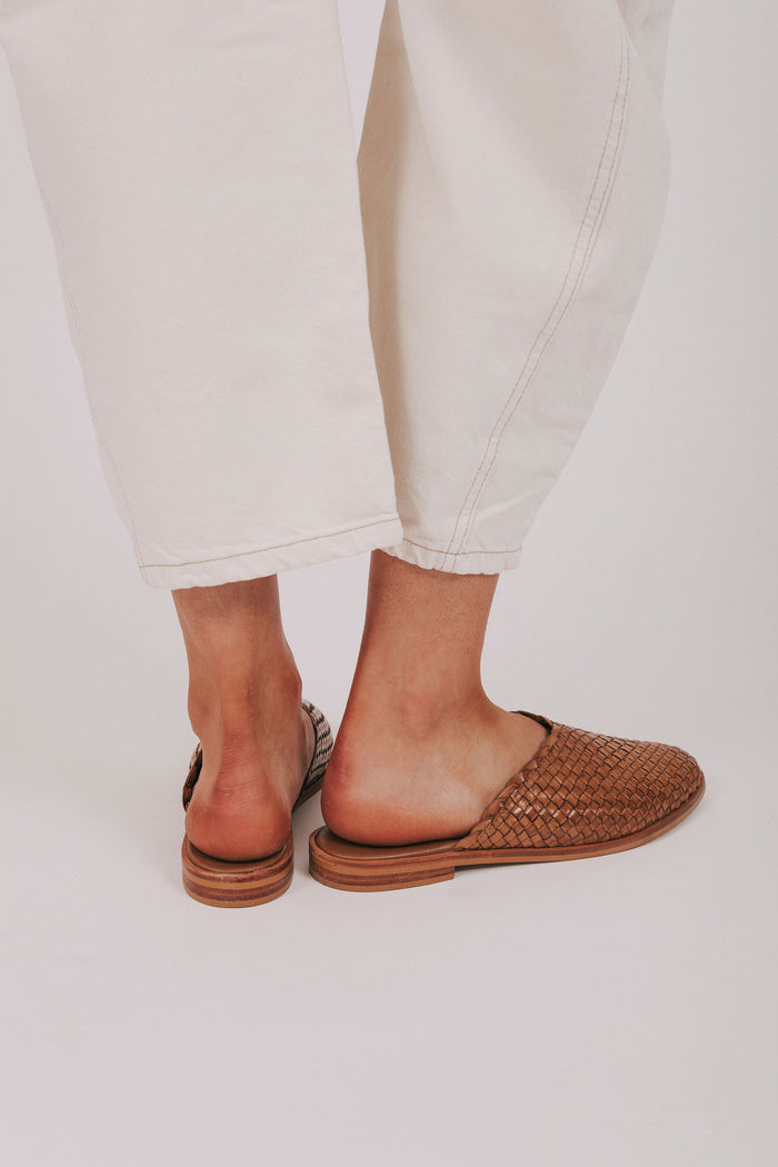 Brown woven sandals worn with light-colored pants on a plain background