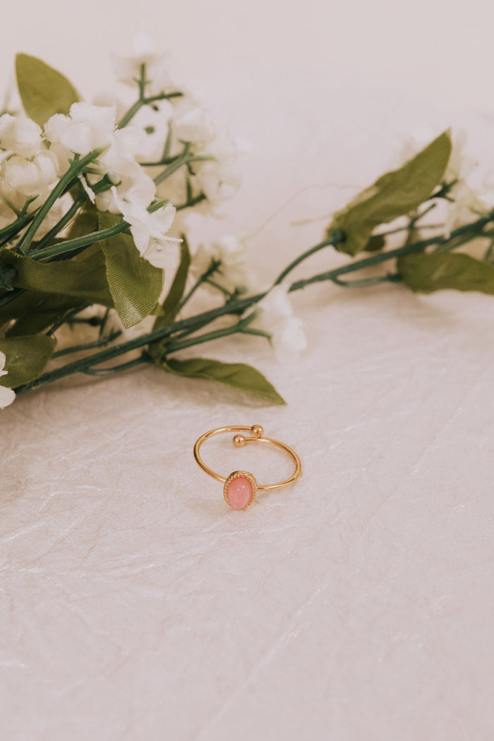 A gold-plated ring with an oval pink stone and textured details, displayed on a white surface with green leaves and white flowers in the background.