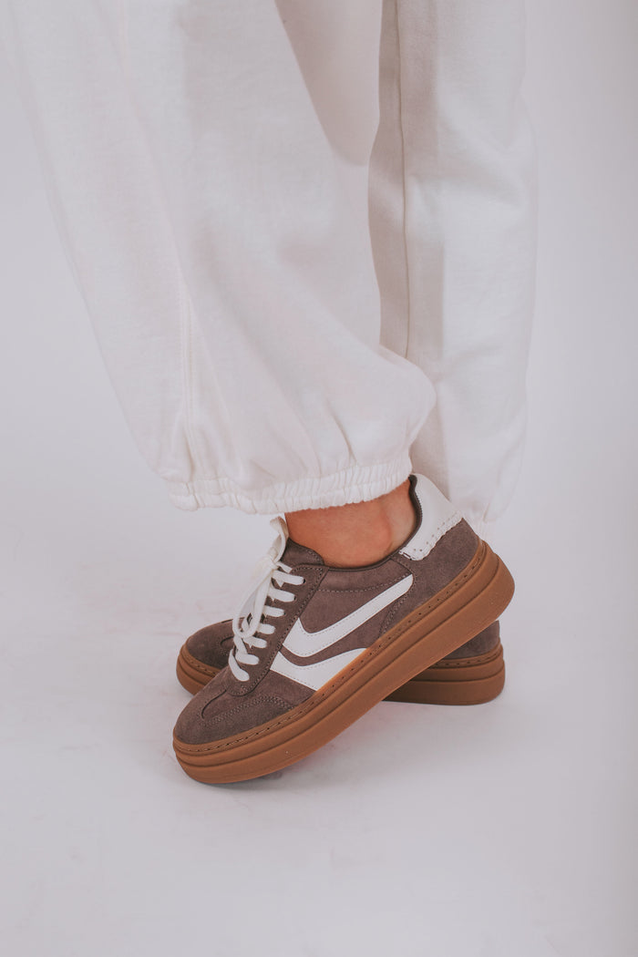 Brown sneakers with white pattern worn with light-colored pants on a white background