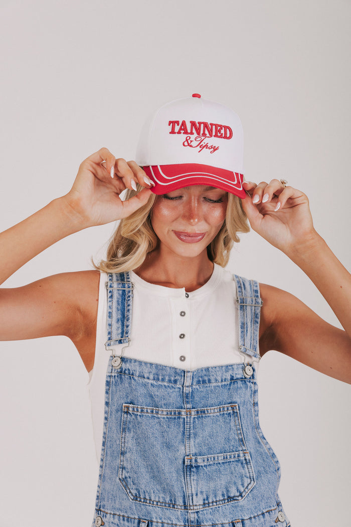 Person wearing a red and white cap with 'Tanned & Tippy' text, white shirt, and denim overalls on a plain background