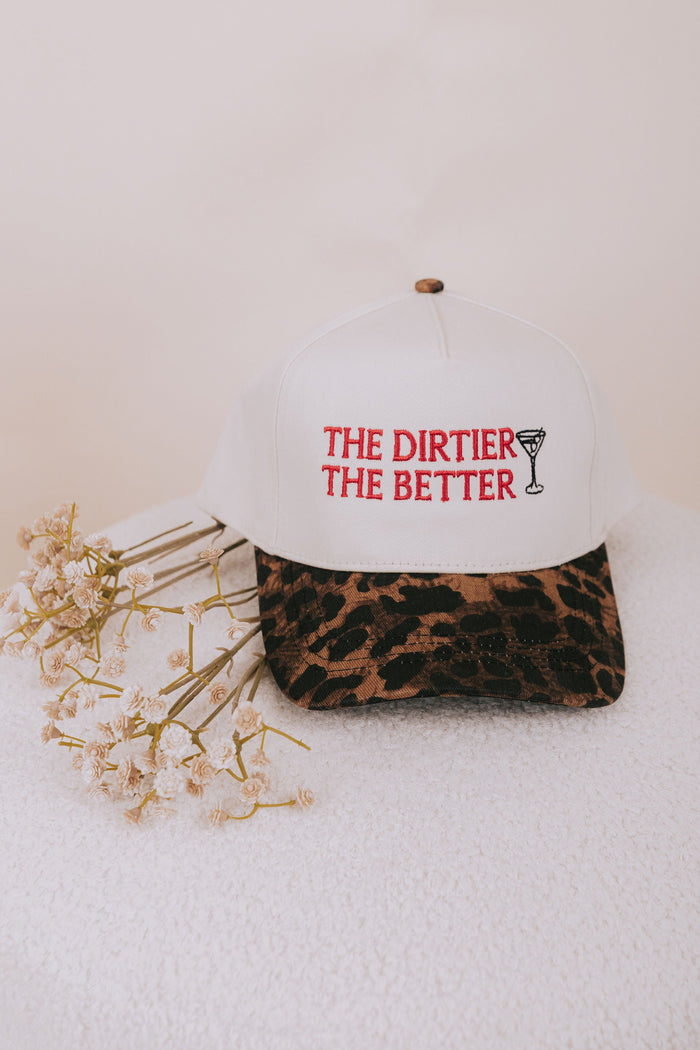White cap with 'The Dirtier The Better' text and leopard print strap on a white surface with flowers.
