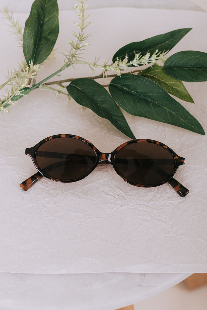 Sunglasses with tortoiseshell frame and dark lenses on a light background with green leaves.
